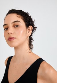 Čierny top bez rukávov s výstrihom do V. Kuceravé vlasy, jemný make-up, zlaté kruhové náušnice a svetlá pleť s pehami. Neutrálny pozadí.