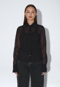 Musta läpinäkyvä pitkähihainen paitapusero, jossa on kaulus ja hienovarainen tekstuuri. Yhdistetty tummiin farkkuihin, joissa on etutaskut.