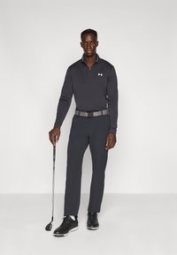 Μαύρο πόλο με μακριά μανίκια και φερμουάρ στον γιακά, συνδυασμένο με σκούρο γκρι παντελόνι και μαύρα παπούτσια γκολφ. Κρατάει ένα μπαστούνι γκολφ και φοράει γκρί ζώνη.