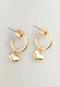 Orecchini a cerchio in tono oro con finitura liscia, dotati di un ciondolo a forma di cuore pendente. La parte hardware include un design con perno e chiusura.