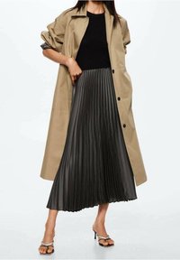 Abrigo trench beige con mangas remangadas, combinado con una falda negra plisada hasta la rodilla en una tela brillante. Sandalias de tacón negras con tiras.