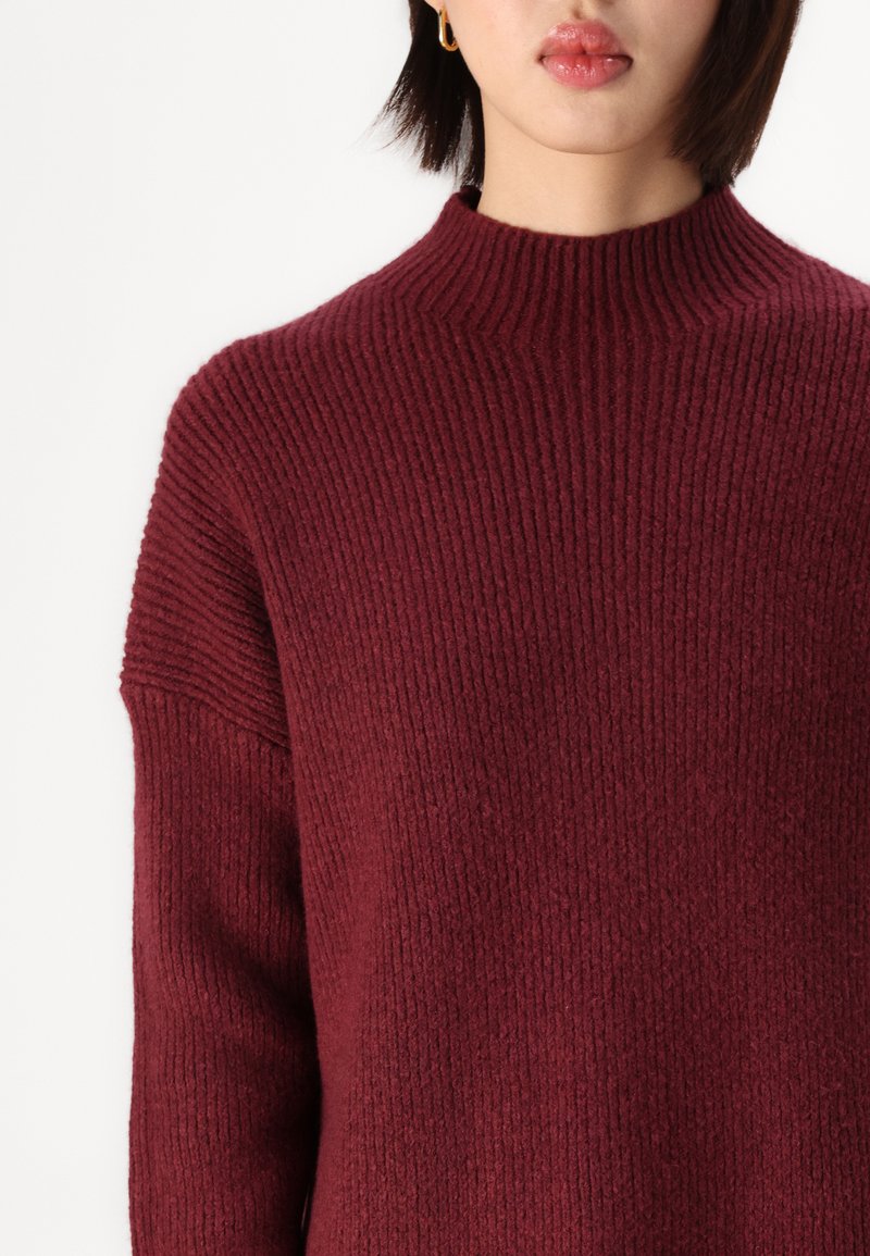 Burgunderfarbener Strickpullover mit hohem Kragen und geripptem Muster. Mit heruntergelassenen Schultern und lockerer Passform, die einen weichen Stoff zur Geltung bringt.