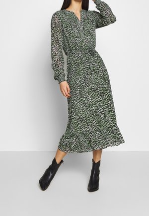 Femme portant une robe midi à manches longues verte et noire à pois, avec une ceinture nouée à la taille et un ourlet à volants, associée à des bottines noires.