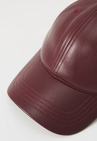 Burgunderfarget skinnlue med en strukturert design, med synlige sømmer og en avrundet skjerm. Glatt tekstur med subtil glans.
