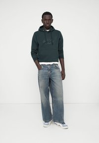 Sweat à capuche vert avec un logo sur le devant, porté sur une chemise blanche. Jean large bleu clair et baskets blanches avec accents bleus. Fond simple.