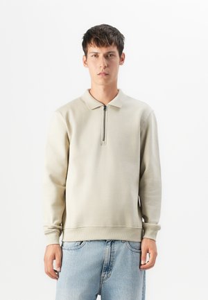 Ung mann iført en beige langermet zip-up kragesweater og lyseblå jeans, stående mot en enkel hvit bakgrunn.