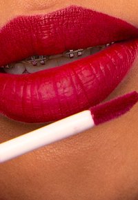 Djup röda läppar med en matt yta, som visar fin textur och definition. En rosa applikator är placerad nära läpparna, vilket förstärker den livfulla färgen.