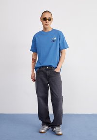 Zils kokvilnas T-krekls ar logo uz krūtīm, kombinēts ar brīviem melniem džinsiem un sudraba sporta apaviem, modelis valkā dzeltenos saulesbrilles.