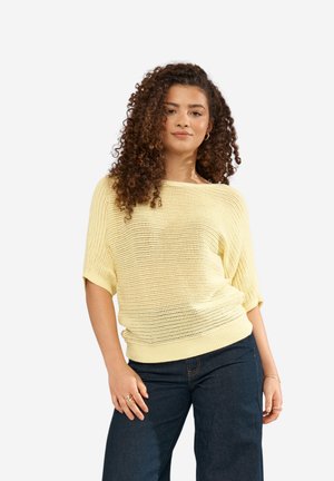 Młoda kobieta o kręconych włosach w luźnym, jasnżółtym swetrze dzianinowym i ciemnoniebieskich dżinsach, stojąca w swobodnej pozie.