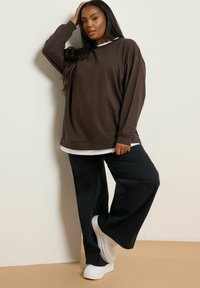 Brauner übergroßer Pullover, der über einem weißen Hemd getragen wird, kombiniert mit schwarzen weiten Hosen und weißen Sneakers. Glattes Material mit lässiger Passform.