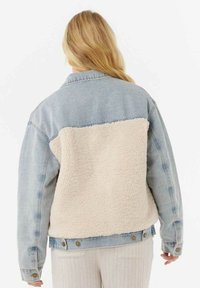 Denimjakke med en lyseblå front og en blød, cremefarvet shearling ryg. Har knaplukninger og et klassisk kravedesign.