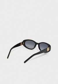 Μαύρα οβάλ γυαλιά ηλίου με σκούρους φακούς και χρυσές λεπτομέρειες στους βραχίονες, τοποθετημένα σε λευκό φόντο.