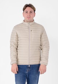 Giacca imbottita beige con motivo a righe orizzontali, chiusura con zip, collo alto e polsini elasticizzati. Indossata sopra jeans blu.