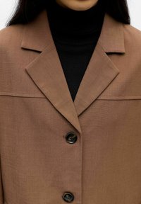 Bruine, op maat gemaakte jas met een kraag met inkeping, zwarte knopen, een gladde textuur en een subtiel naden detail over de borst.