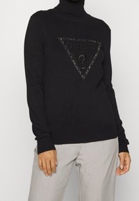 Maglione dolcevita nero con una toppa triangolare in rilievo con la scritta "GUESS" e decorazioni ornamentali. Indossato con pantaloni grigio chiaro.