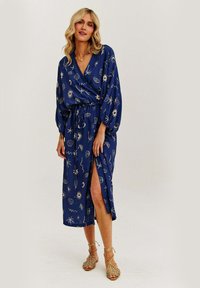 Modré balonové šaty s potiskem ve zlatém plážovém stylu, s dlouhými balónovými rukávy, pásem v pase a rozparkem až k stehnu. Hladká textilie.