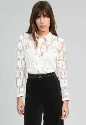 Femme aux cheveux bruns lisses portant un chemisier transparent en dentelle blanche avec des boutons et un pantalon en velours noir taille haute, posant devant un fond gris.