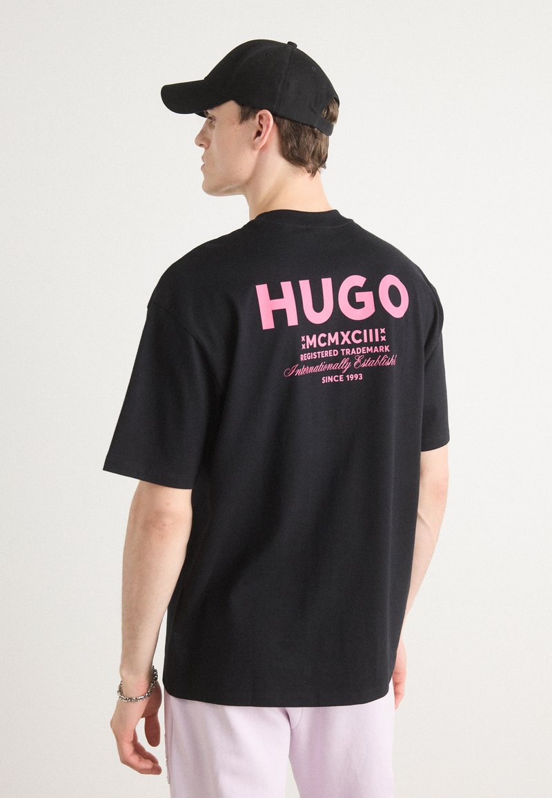Fekete pamut póló, nagyméretű rózsaszín "HUGO" felirattal a hátsó részen, további márkázási elemekkel és laza szabással.