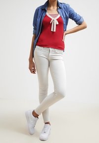 Persona vestida con una blusa roja con un lazo, camisa de mezclilla azul, pantalones skinny blancos y zapatillas blancas, de pie con una mano en la cadera.