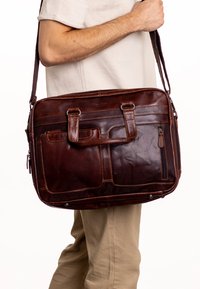 Borsa a tracolla in pelle marrone con due manici superiori e una lunga tracolla. Presenta diverse tasche e dettagli di cucitura. Texture liscia.