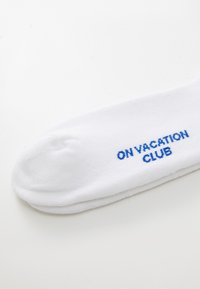 Bela nogavica iz prepustljive tkanine z modro izvezenim napisom "ON VACATION CLUB" na strani. Gladka tekstura in okrogla oblika prsta.