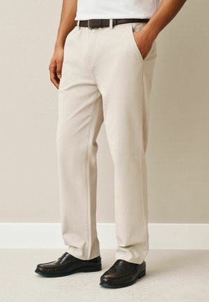 Mann, der beige Hose, schwarzen Gürtel, weißes Hemd und dunkelbraune Loafers trägt, steht mit einer Hand in der Tasche vor einem einfarbigen Hintergrund.