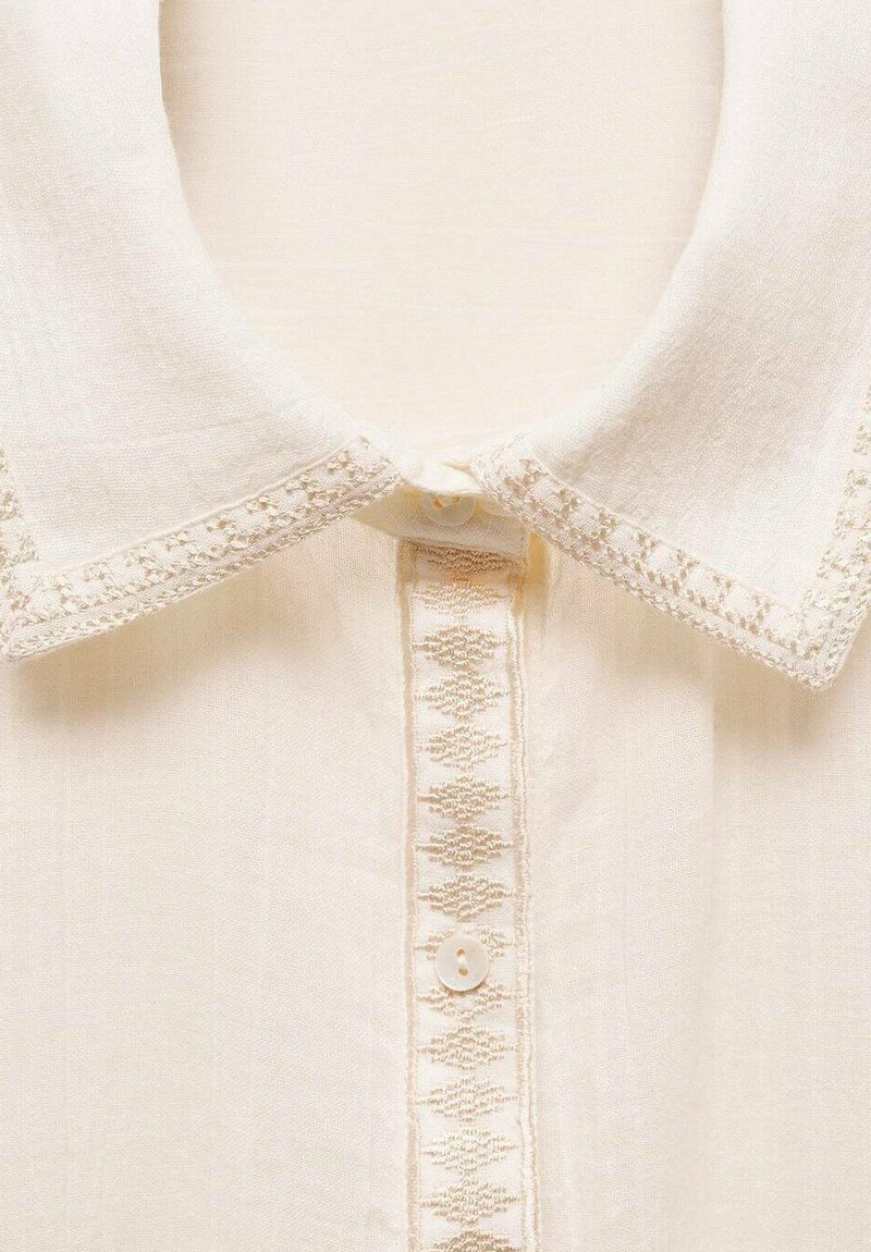 Blusa color crema con colletto, caratterizzata da un sottile bordo ricamato lungo il colletto e la lista dei bottoni, con chiusura a un solo bottone. Tessuto morbido e testurizzato.