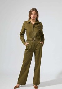 Combinaison vert olive en tissu texturé ; comporte un devant à boutons, des manches longues, un col, une ceinture à la taille avec boucle ronde et des jambes larges.