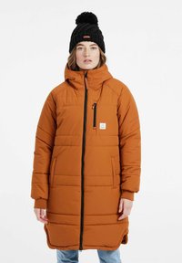 Polstret orange lang frakke med høj krave, frontlynlås, sidelommer og logo-patch. Sort strikket hat med pom-pom detalje på toppen.