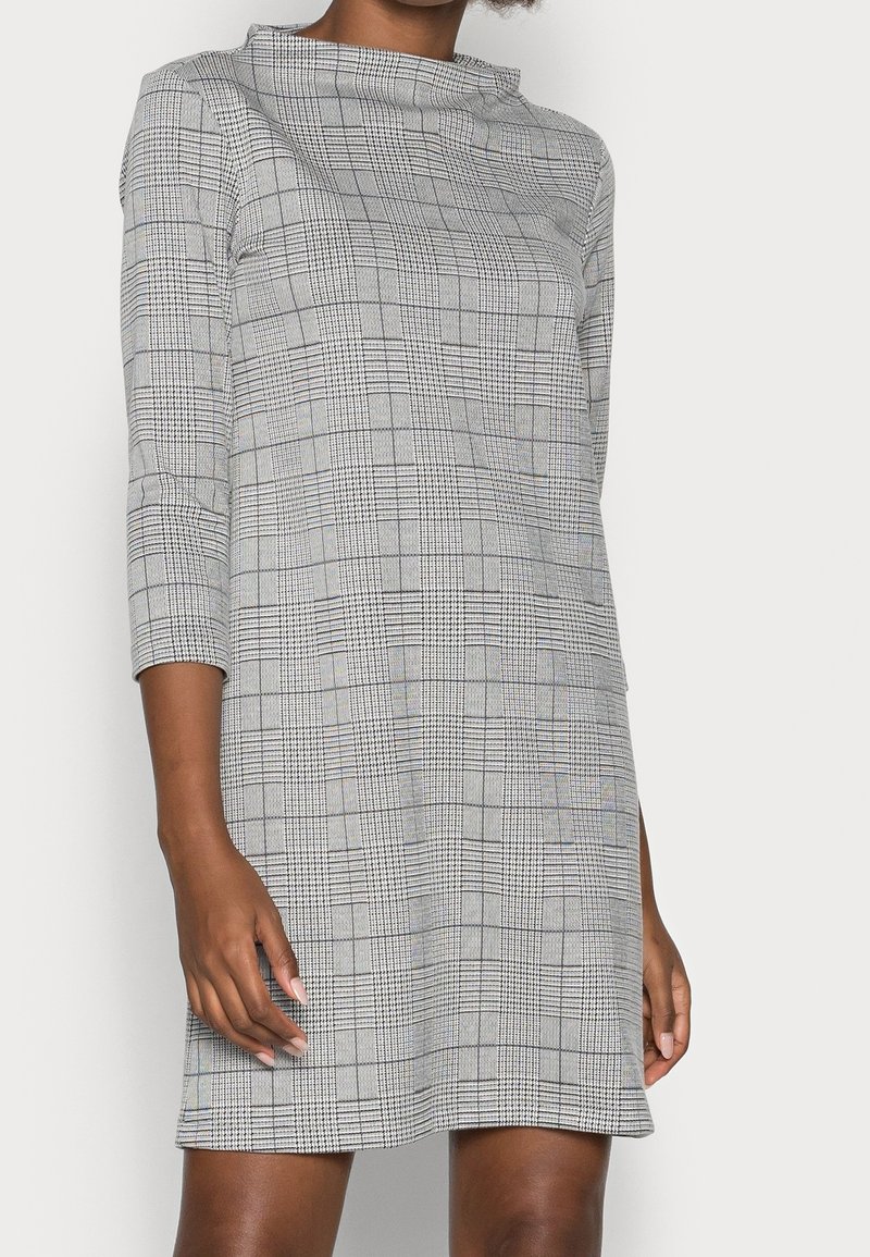 Femme portant une robe à carreaux grise avec des manches trois-quarts et un col haut, debout devant un fond clair uni.