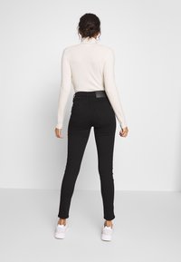 Mustat kapeat farkut, joissa on korkea vyötärö ja takataskut, yhdistettynä kermanväriseen pitkähihaiseen paitaan. Mallilla on valkoiset lenkkarit.