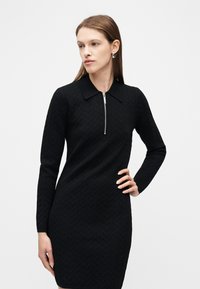 Γυναίκα που φοράει μαύρο στενό φόρεμα με μακριά μανίκια και γιακά, με υφή ζιγκ-ζαγκ και ασημένιο ημικυκλικό φερμουάρ, ποζάρει με το ένα χέρι στη μέση.