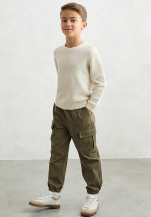 Krémový pletený sveter s rebrovanými manžetami, olivové cargo nohavice s bočnými vreckami a biele tenisky s béžovými doplnkami. Celková hladká textúra.