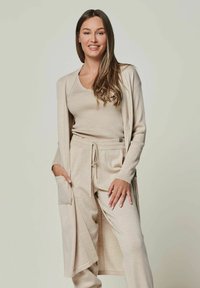 Beige Strick-Loungewear-Set mit einem langen Cardigan mit Taschen, einem Rundhalsausschnitt-Oberteil und Jogginghosen mit Kordelzug, alles aus einem weichen, strukturierten Stoff.