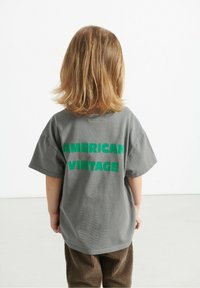 Hallitus tooniga T-särk lühikeste varrukatega, mille tagaküljel on paksud rohelised tähed sõnumiga "AMERICAN VINTAGE". Kombineeritud pruunide sametpükstega.