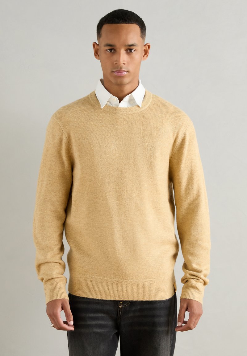 Helles beige Sweatshirt mit Rundhalsausschnitt, langen Ärmeln und gerippten Bündchen und Saum. Über einem weißen Kragenhemd getragen, in lässiger Passform. Keine sichtbaren Muster.