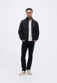 Μαύρο bomber μπουφάν με ψηλό γιακά, φορεμένο πάνω από λευκό πόλο. Μαύρο τζιν παντελόνι και μπεζ αθλητικά παπούτσια ολοκληρώνουν το σύνολο.