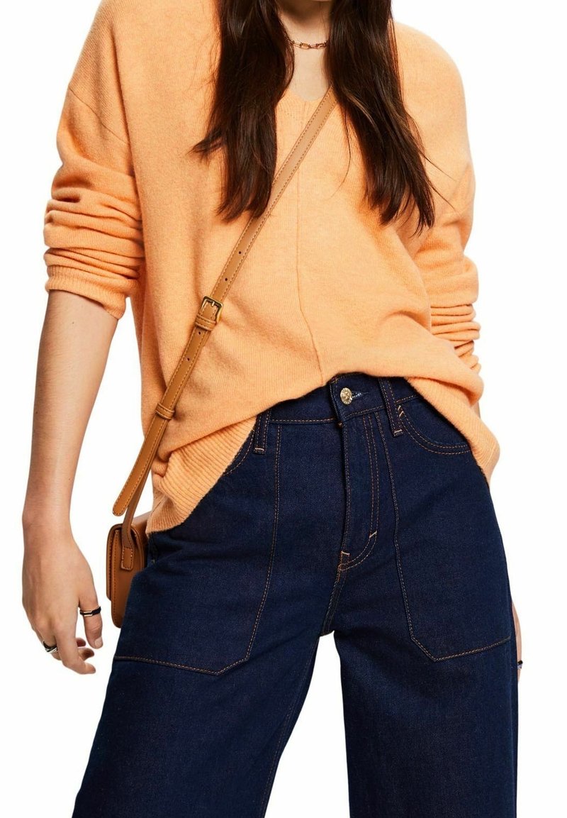 Pomarańczowy sweter dzianinowy z podwiniętymi rękawami w połączeniu z wysokim stanem ciemnoniebieskich dżinsów, które mają złoty guzik i kontrastowe przeszycia.