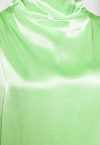 Detailní záběr na hladkou, lesklou, světle zelenou látku elegantně zavěšenou a předvádějící svou texturu a lesk na neutrálním pozadí.