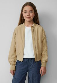 Veste bomber beige en tissu lisse avec une fermeture éclair à l'avant, des poignets côtelés et une poche sur le bras gauche. Portée sur une chemise blanche.