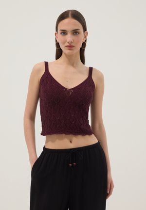 Joven con cabello castaño liso lleva pendientes de aro, un crop top de punto sin mangas de color púrpura oscuro y pantalones negros holgados con cordón.