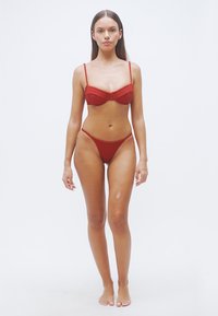 Σετ σουτιέν και μπικίνι σε σκουρόκοκκινο, με σχεδιασμό από υφή με ανάγλυφες ρίγες. Το σουτιέν διαθέτει σύστημα υποστήριξης με μπανέλα, ενώ το κάτω μέρος του μπικίνι έχει ψηλωμένο κόψιμο στα πόδια.
