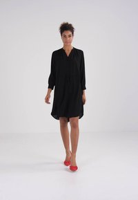 Svart knelång klänning med v-ringning, trekvartsärmar, dragsko i midjan och mjukt tyg. Modellen har på sig röda ballerinas.