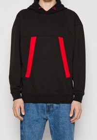 Felpa nera con cappuccio e tasca a marsupio, caratterizzata da strisce verticali rosso vivo. Realizzata in tessuto morbido, ha un cappuccio con coulisse e polsini a coste.