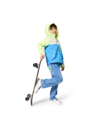 Niño con chaqueta azul y verde sosteniendo una patineta detrás de una pierna, vistiendo jeans y zapatillas blancas, mirando hacia arriba.