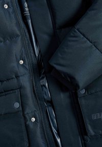 Giacca imbottita blu navy con texture liscia, zip anteriore, bottoni a scatto e polsini a coste, con foderatura interna visibile alla zip.