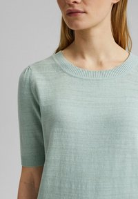 Femme portant un pull léger en maille vert clair à manches courtes, avec un col rond côtelé et un détail bouffant aux épaules.