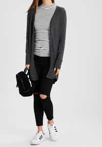 Cárdigan gris abierto sobre una camisa de manga larga a rayas, combinado con jeans ajustados negros rotos y zapatillas blancas. Mochila negra de material peludo.