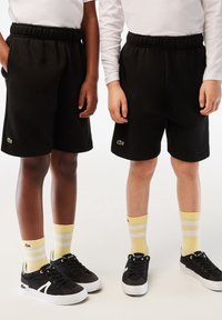 Mustat fleece-shortsit, joissa on joustava vyötärö, pieni vihreä logo, keltaisia raidallisia sukkia ja mustia lenkkikenkiä.