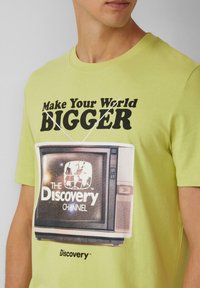 Hellgrünes Baumwoll-T-Shirt mit einer Vintage-Fernsehgrafik und dem Text "Mach deine Welt GRÖßER" sowie "The Discovery Channel" in Schwarz.